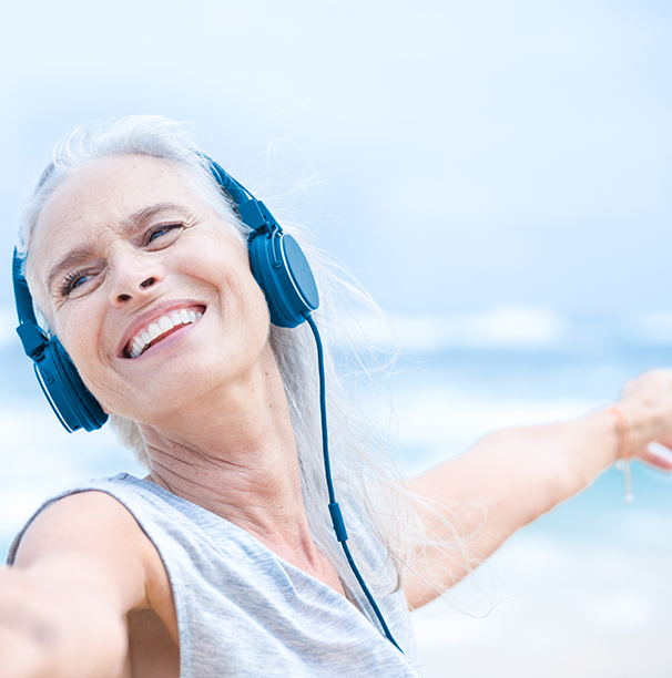 Woman in tank top with ear phones stretching her arm out. She is by the sea.